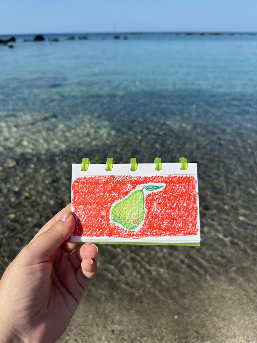 A hand holding a pastel drawing of a green pear on a red background, sea on the background